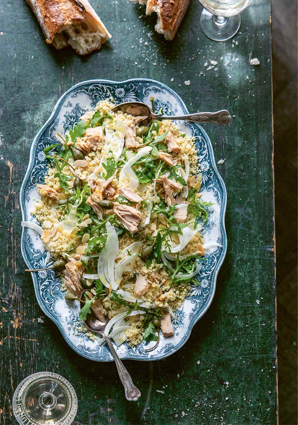 A blue and white platter filled with couscous salad with fennel and almonds with a torn baguette and a couple of glasses of white wine beside it.