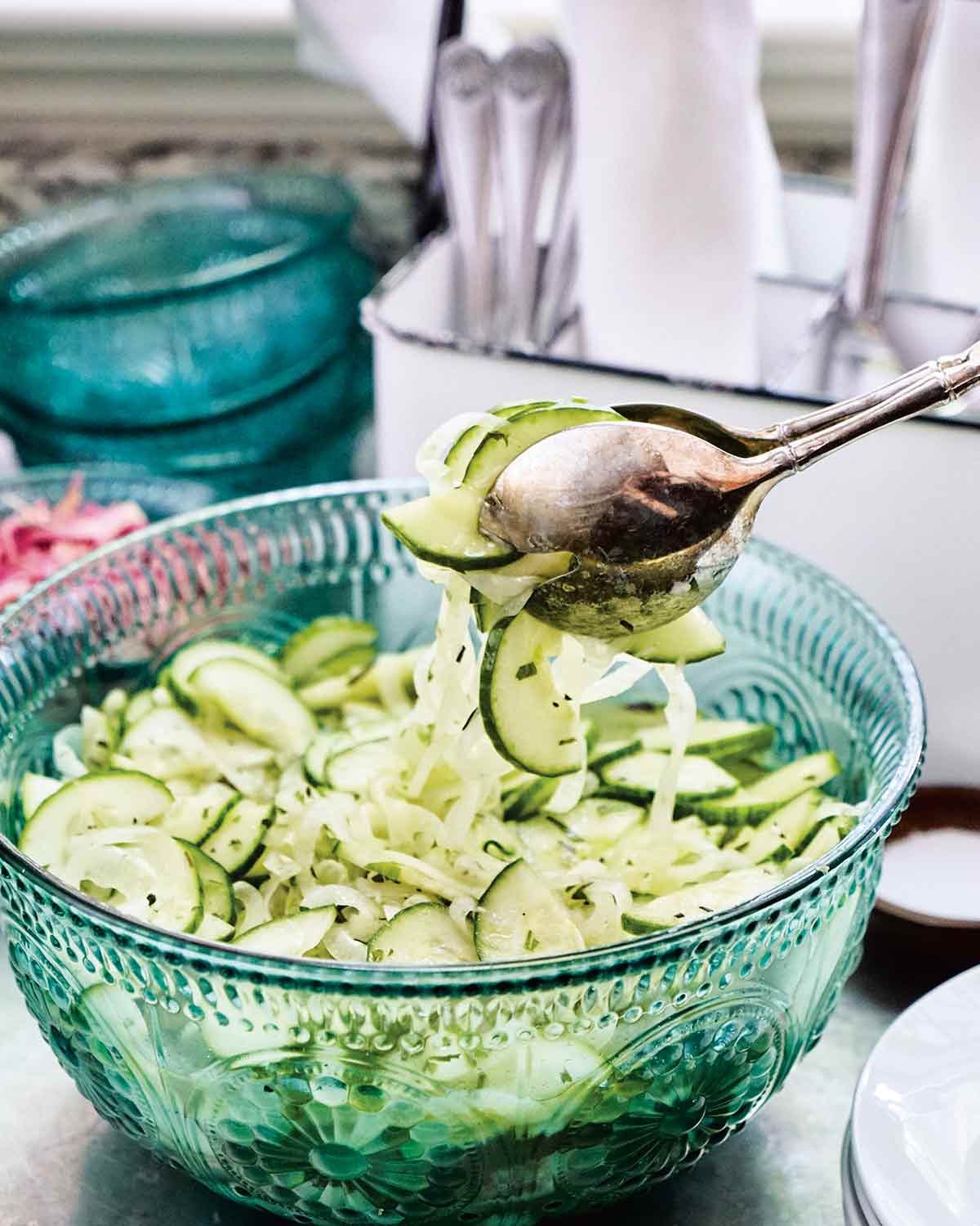 A serving of cucumber and onion salad being lifted from a serving bowl with silver salad tongs.
