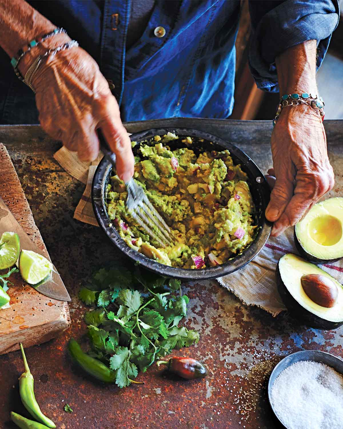 A person mashing avocado into a bowl of easy guacamole on a table with salt, avocado, cilantro, lime, and chiles.