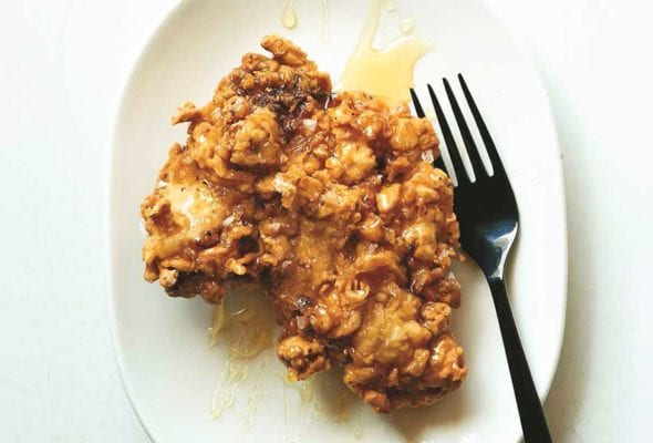 A piece of egg shop fried chicken on a white plate with a drizzle of honey on top and a fork beside it.