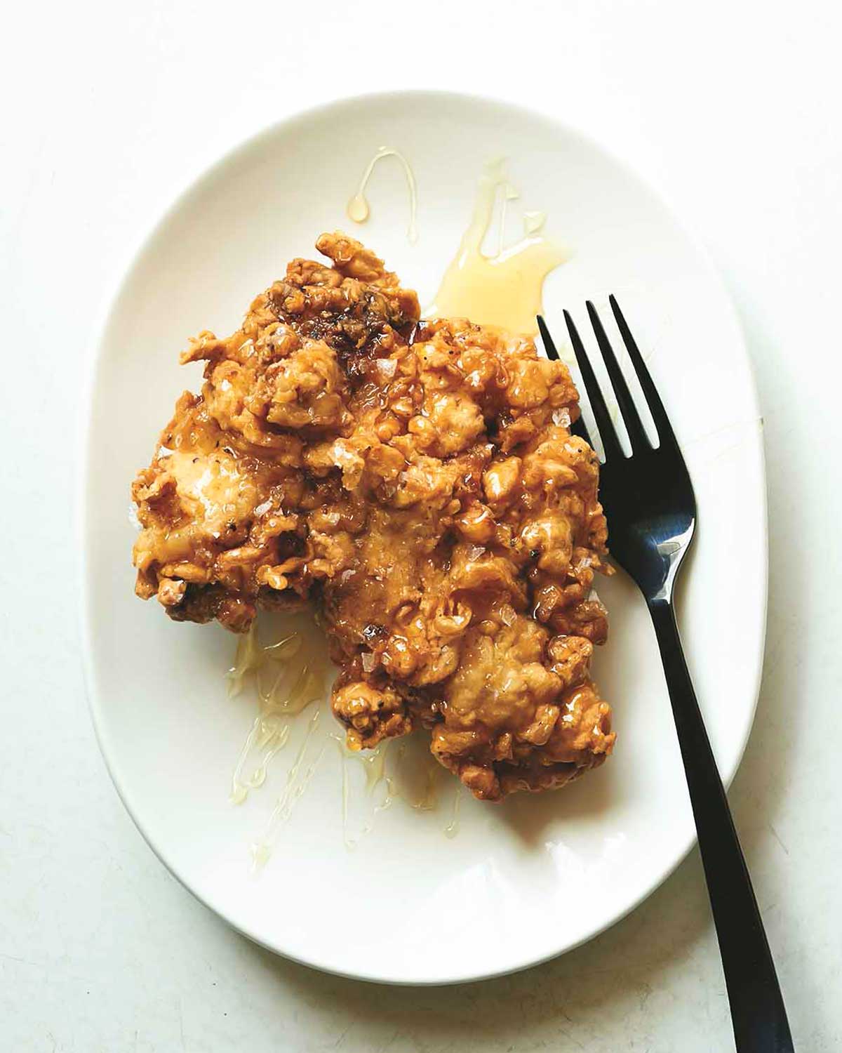 A piece of egg shop fried chicken on a white plate with a drizzle of honey on top and a fork beside it.