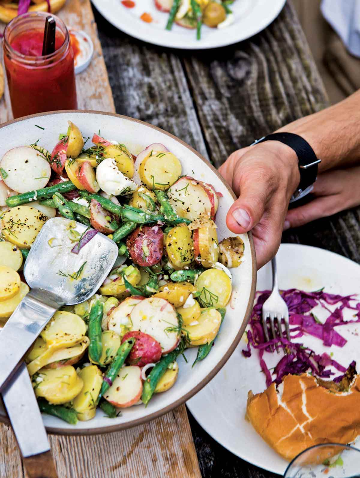A person holding a platter of French potato salad above a wooden table.
