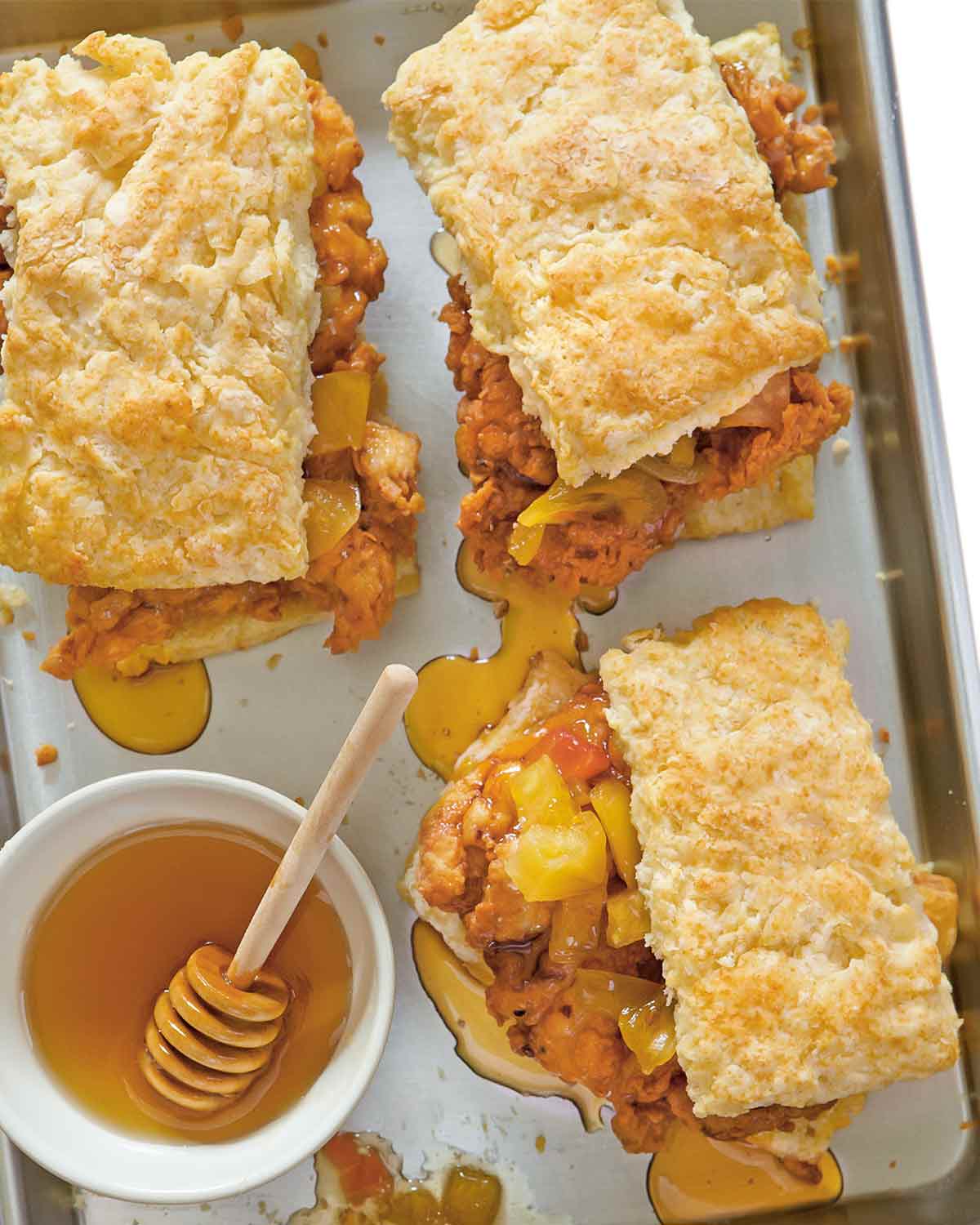 Three fried chicken and biscuits sandwiches on a baking sheet with a bowl of honey beside and drizzled around the sandwiches.
