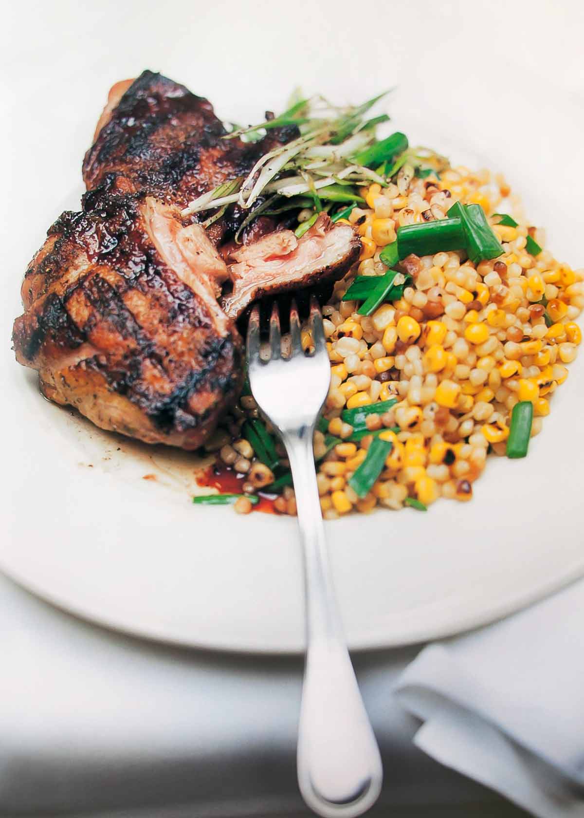 A piece of grilled guinea hen with sweet corn fregula beside it, all topped with chopped scallions.