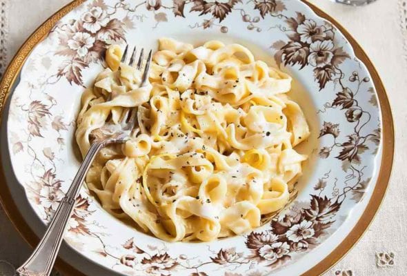 A patterned bowl filled with lemony fettuccine alfredo on a white and gold plate with a glass of water and dish of Parmesan on the side.