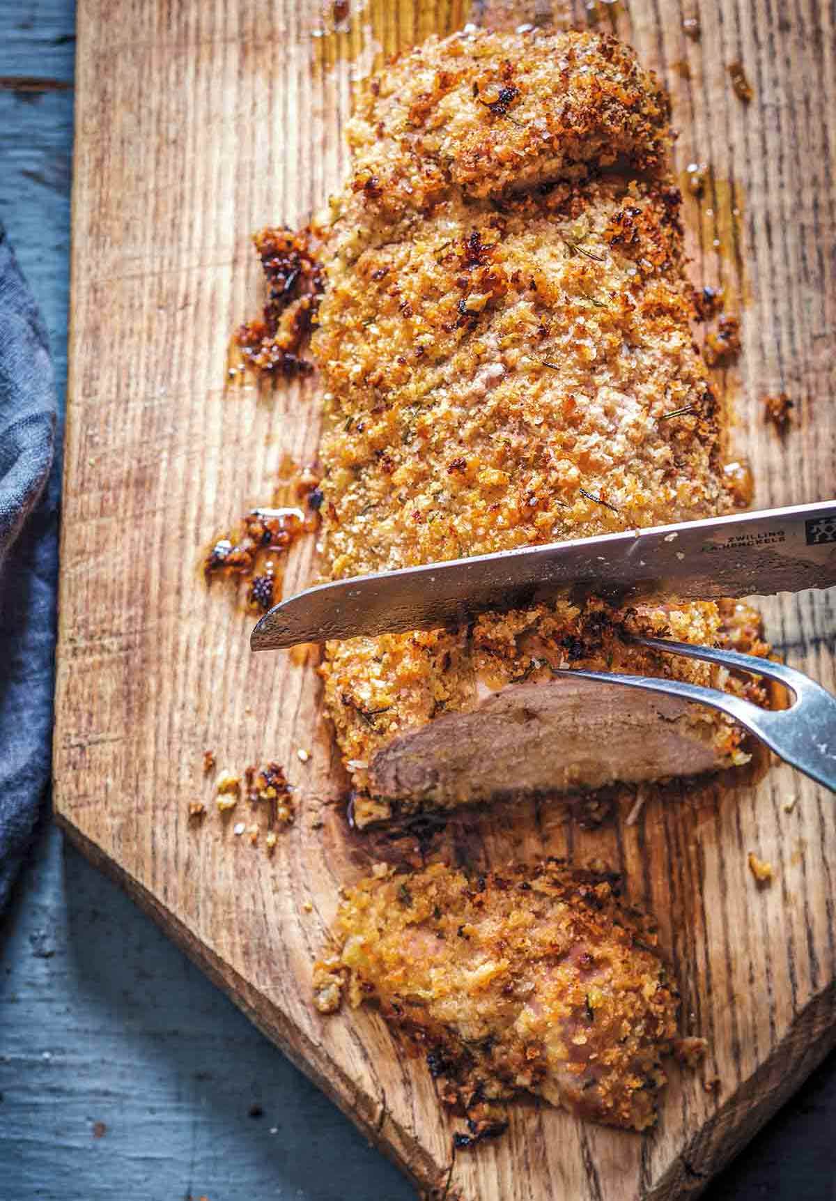 A panko crusted pork tenderloin with rosemary being sliced on a wooden cutting board.