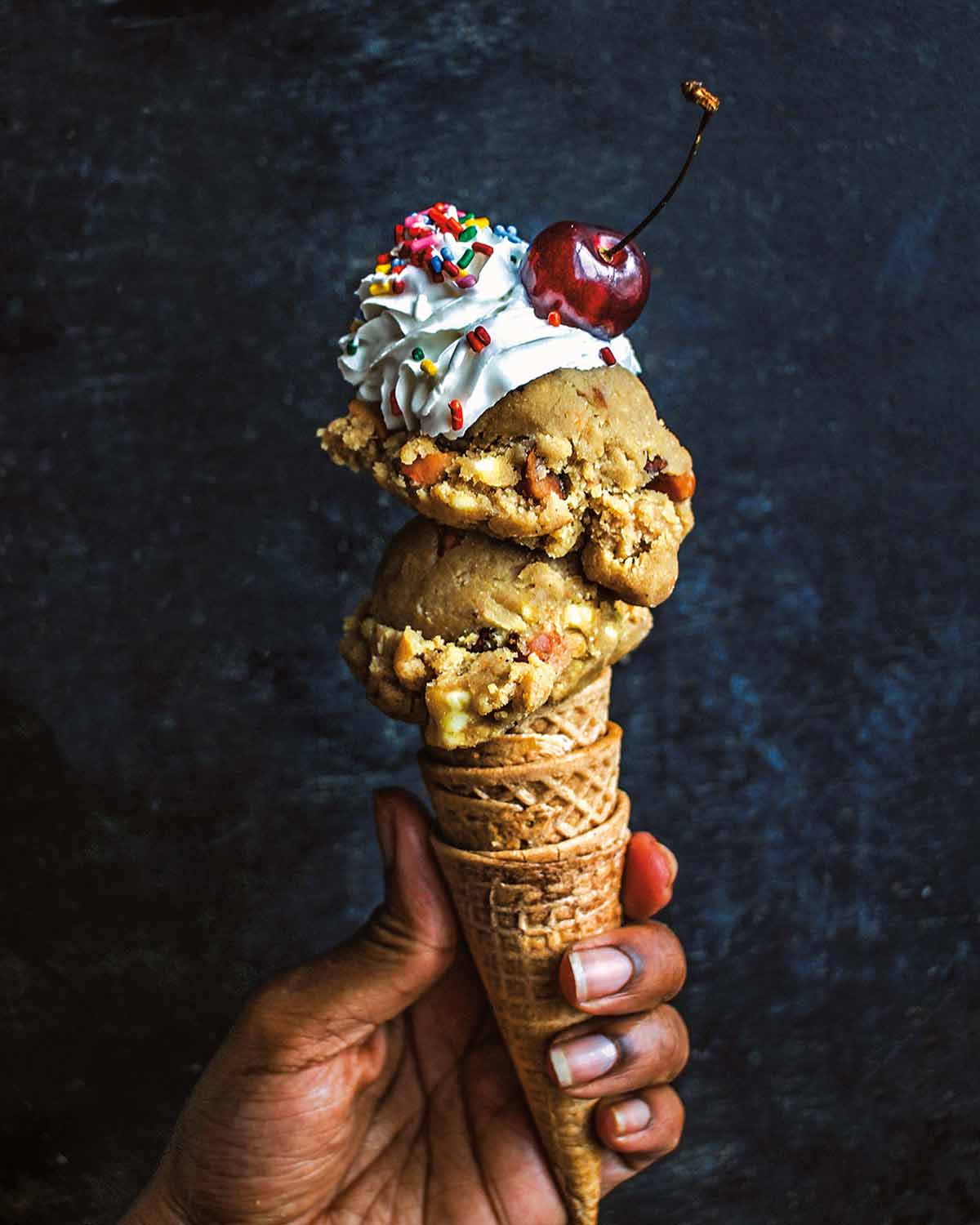 A person holding three nested ice cream cones topped with two scoops of vegan gluten-free chocolate chip cookie dough, whipped cream, sprinkles, and a cherry.