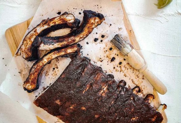 A rack of Vietnamese pork ribs on a sheet of parchment on a cutting board with a basting brush and a bowl of lime wedges nearby.