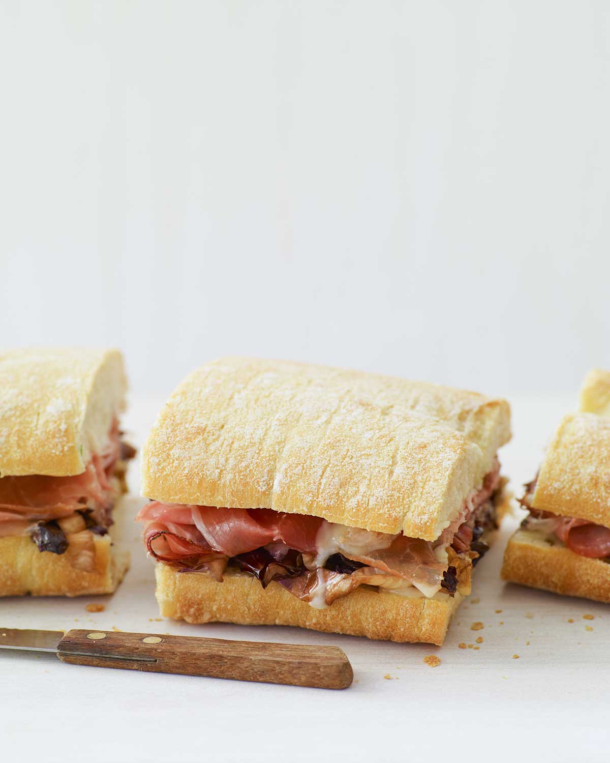 A warm panini with taleggio, grilled radicchio, and speck sandwich cut into thirds with a knife in front of the sandwich.