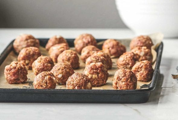 A tray of unbaked bbq meatballs with a set of measuring spoons beside it and a bowl of barbecue sauce in the background.