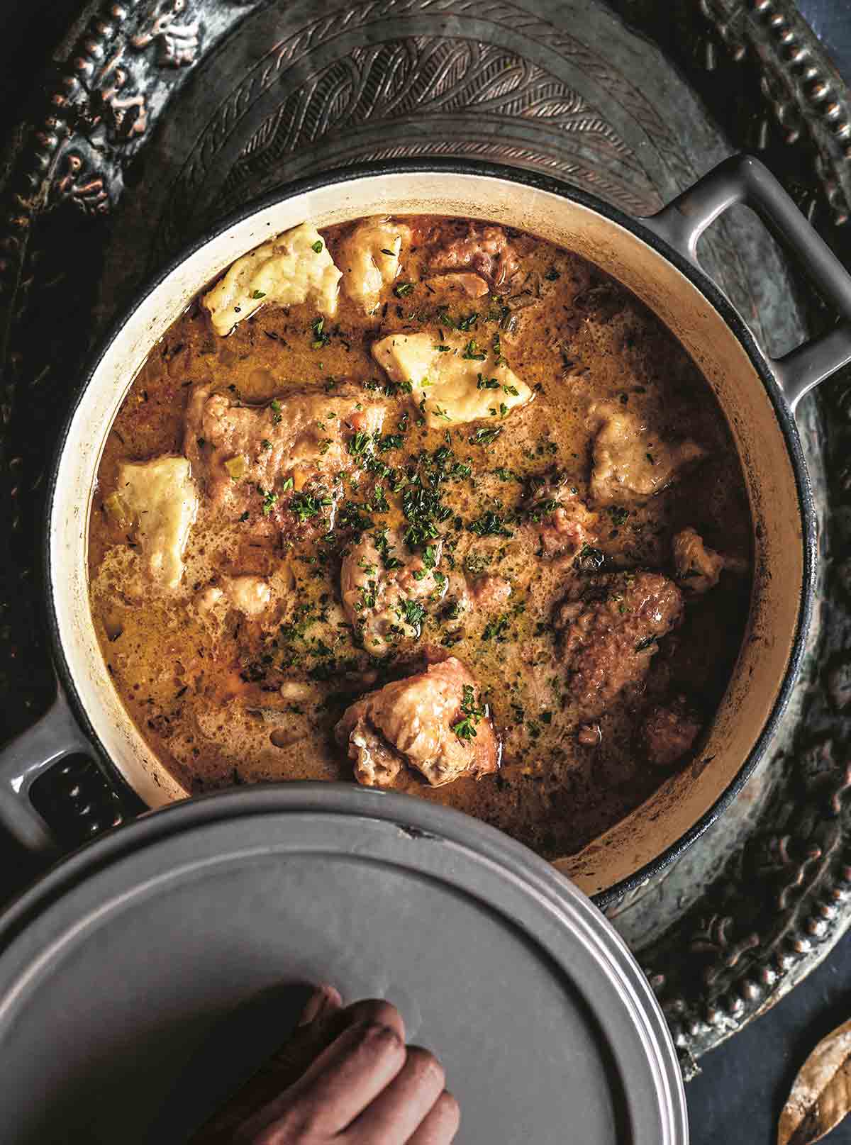 A person lifting the lid off a pot filled with chicken and dumplings.