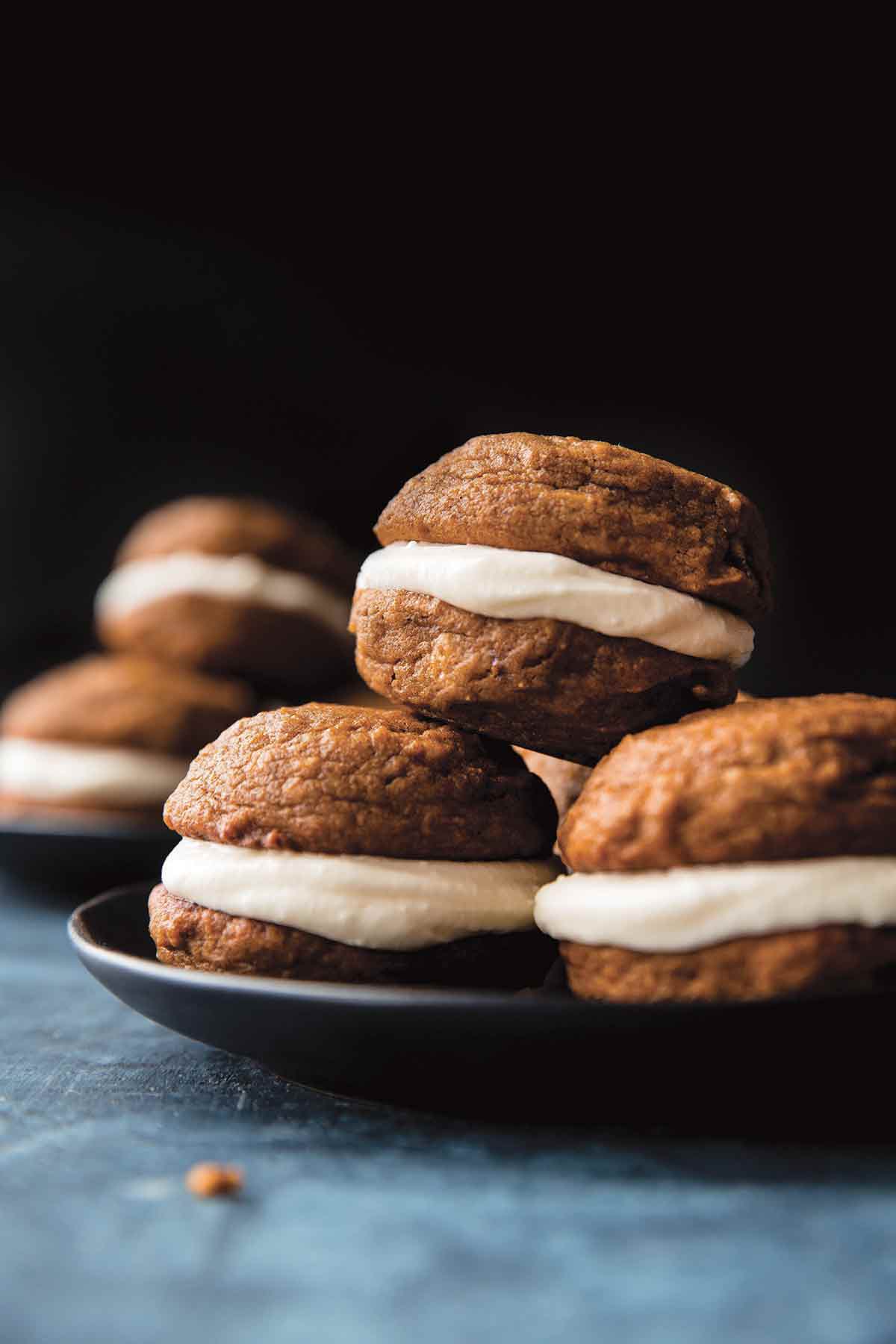 Two plates each with four pumpkin spice whoopie pies with cream cheese filling stacked on them.