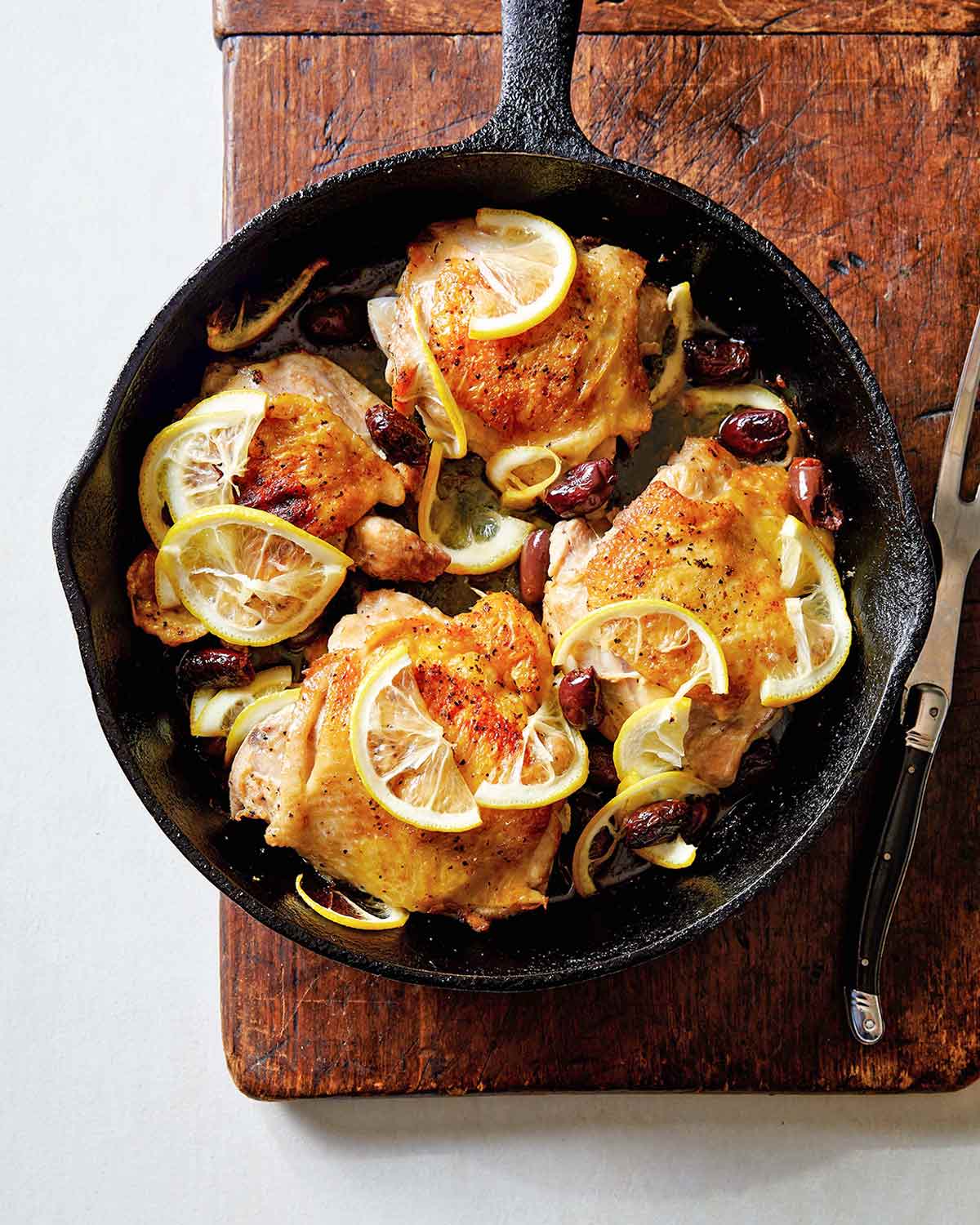 Four roast chicken thighs with lemon in a cast-iron skillet set on a wooden board with a meat fork resting beside it.