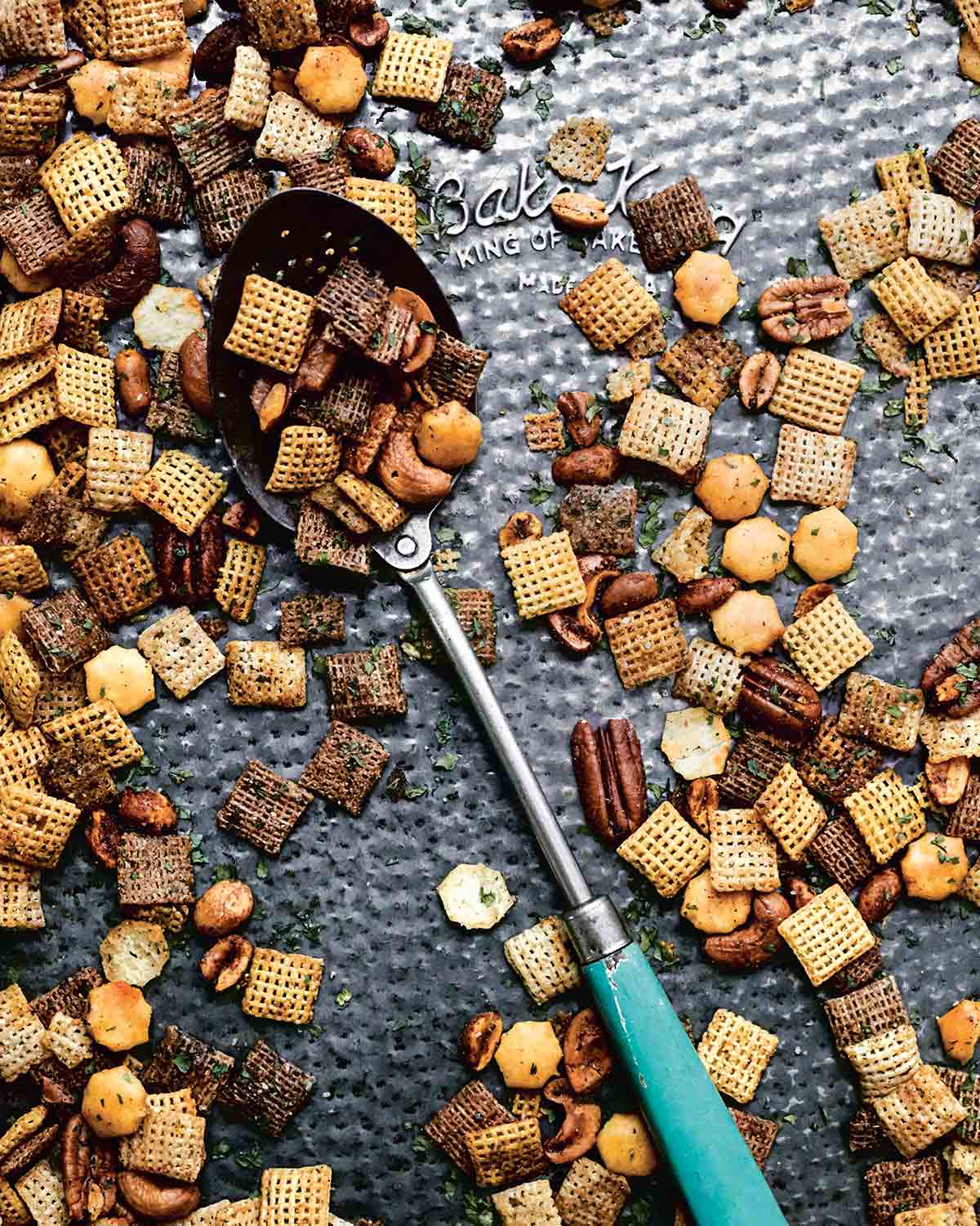 Spicy Chex mix scattered on a baking sheet with a spoon lying in the middle.