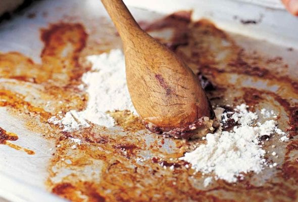 A person stirring flour into the basic pan gravy with a wooden spoon on a rimmed baking sheet.