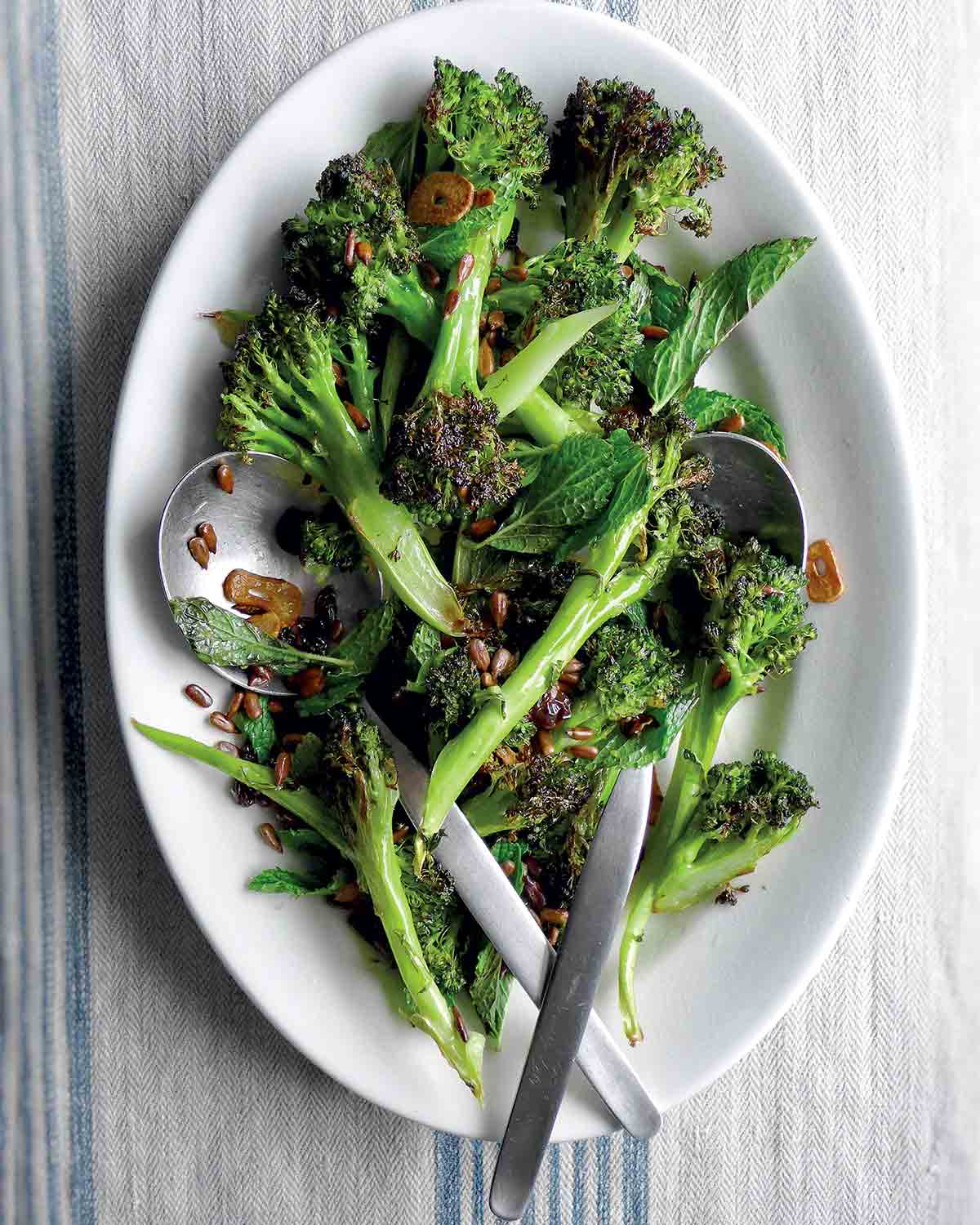 A white plate with spears of roasted broccoli with pickled golden raisins scattered overtop and two spoons in the broccoli.