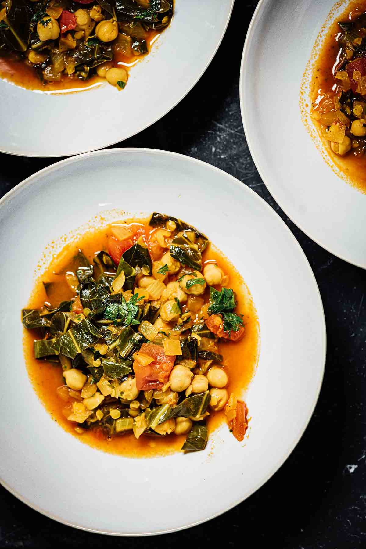 Three white bowls filled with collard greens, chickpea, and lentil soup.
