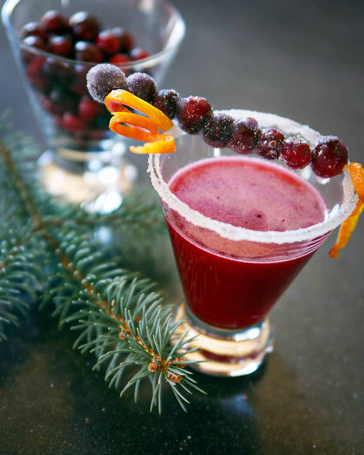 A cranberry margarita in a short glass with a skewer or sugared cranberries and orange zest across the top.