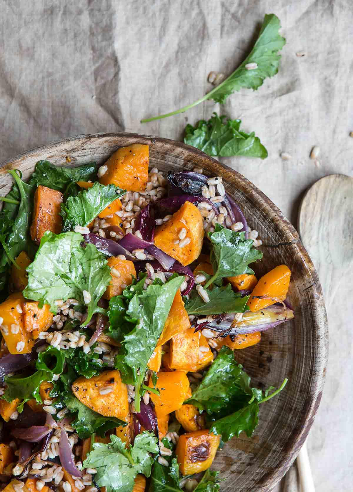A wooden bowl filled with kale butternut squash farro salad with a wooden spoon resting beside the bowl.