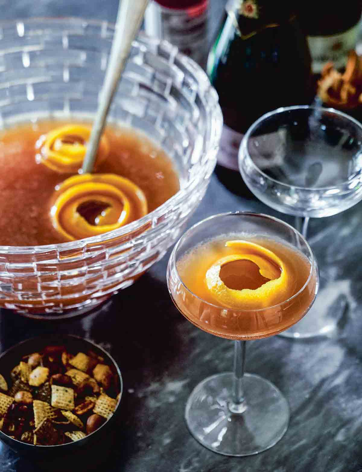 A glass punch bowl partially filled with maybelle punch and orange peel garnish, and two coupe glasses alongside, one filled and one empty.