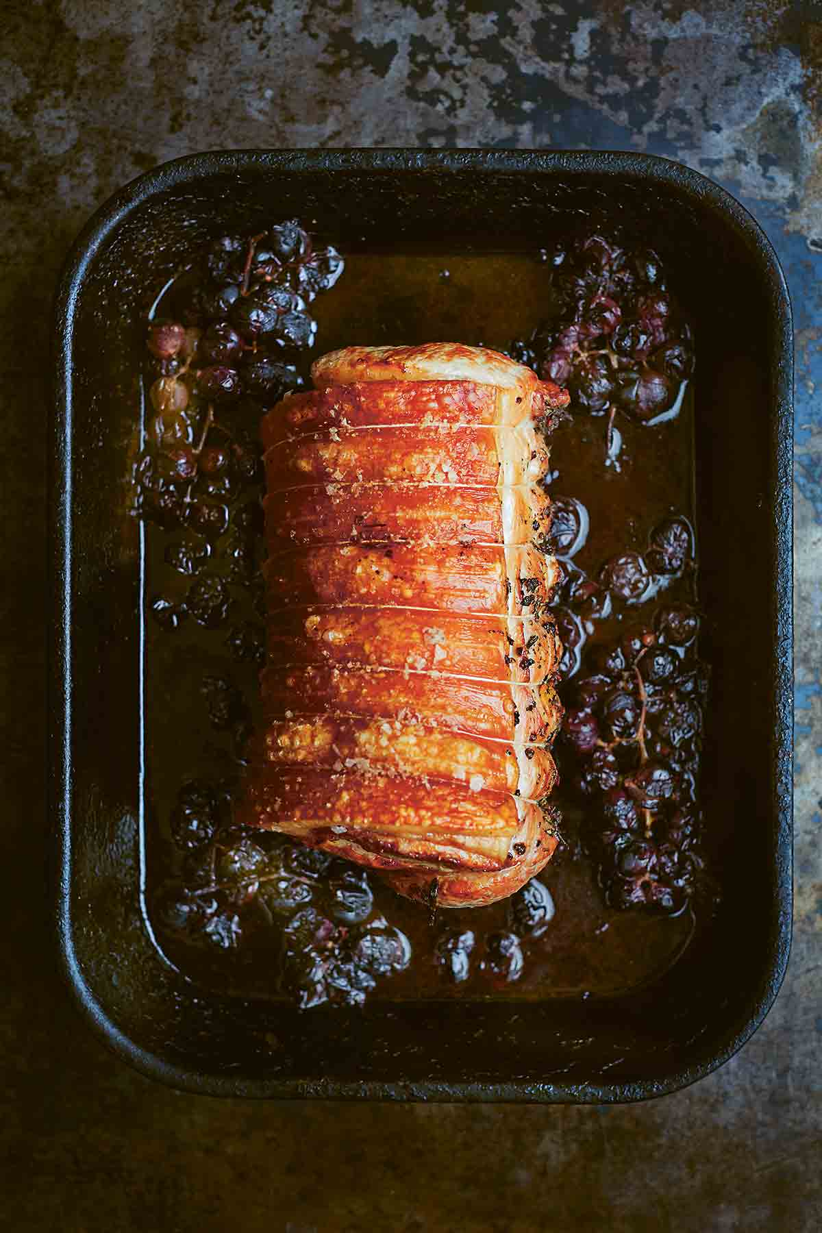 A tied roast pork with crushed grapes in a roasting pan.