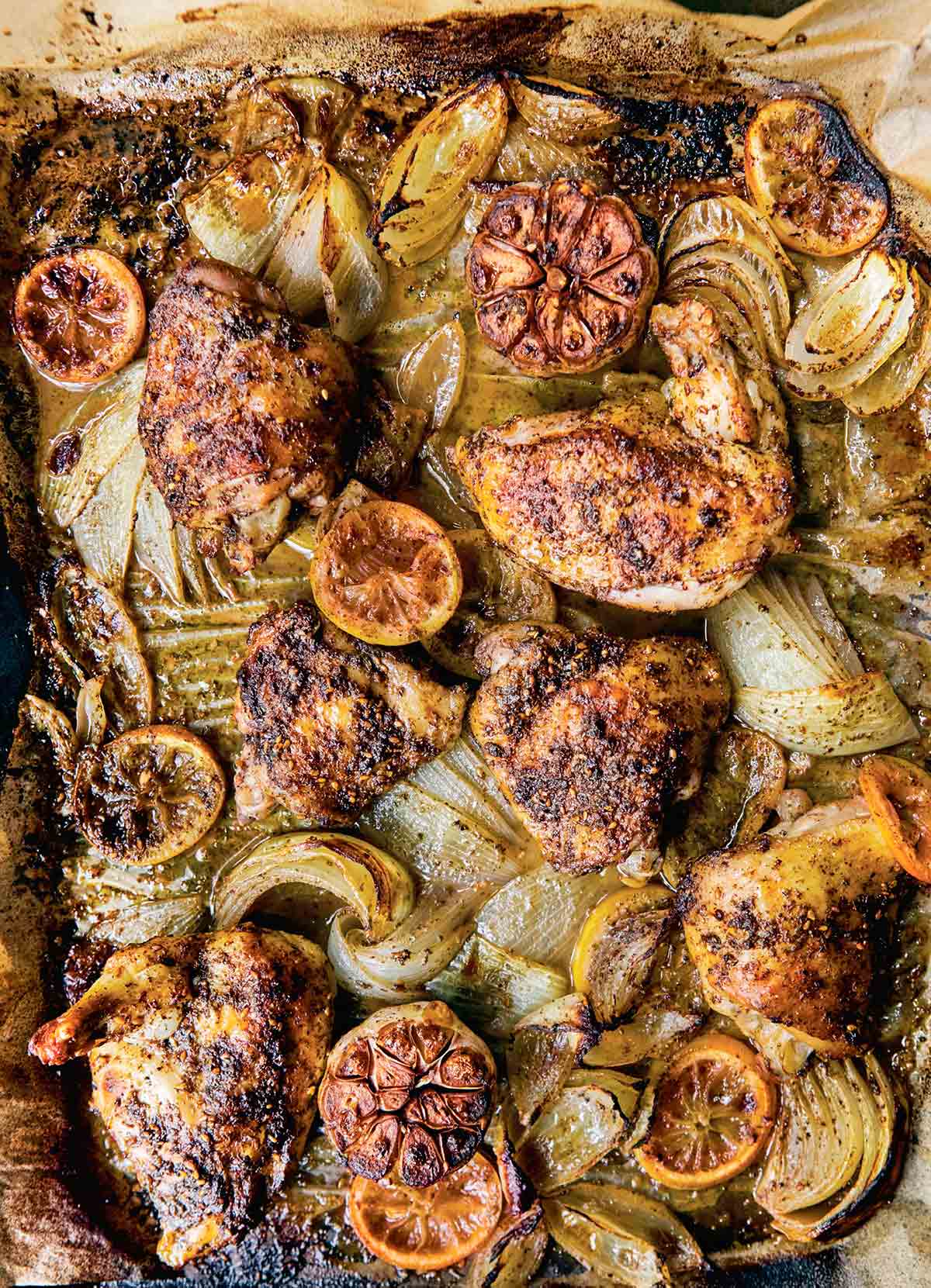Pieces of sheet pan roast chicken with za'atar with onion wedges, halved garlic heads, and lemon slices.