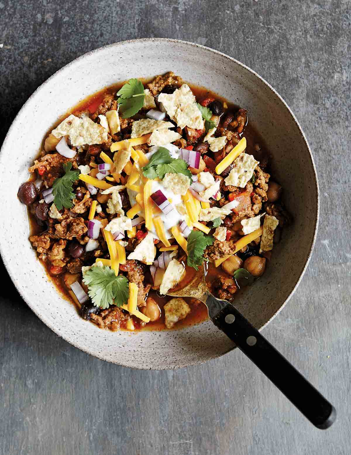 An earthenware bowl filled with slow cooker beef chili, topped with cheese, tortilla chips, and cilantro, with a spoon resting in it.