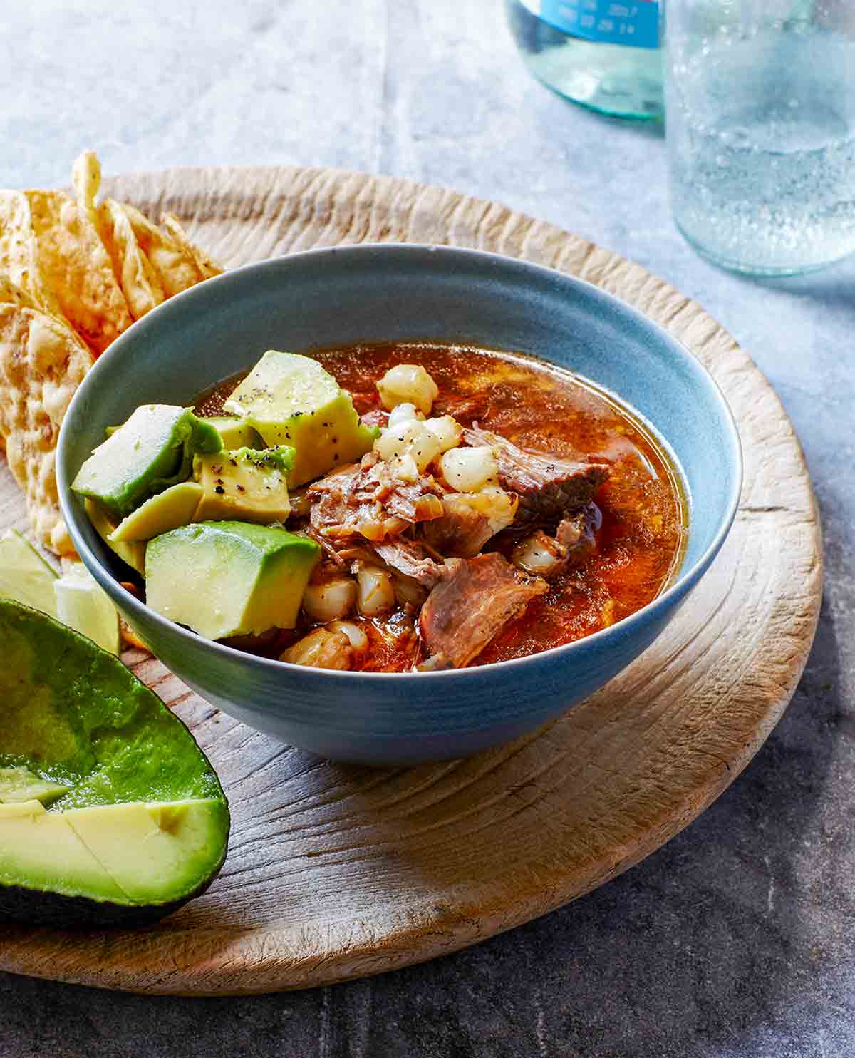 A bowl of pork, avocado, and hominy in a red broth on a cutting board with tortilla chips