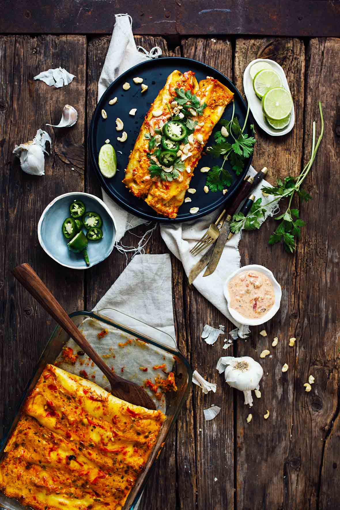 A casserole dish of sweet potato enchiladas with a place with two enchiladas, lime, jalapenos, and sauce