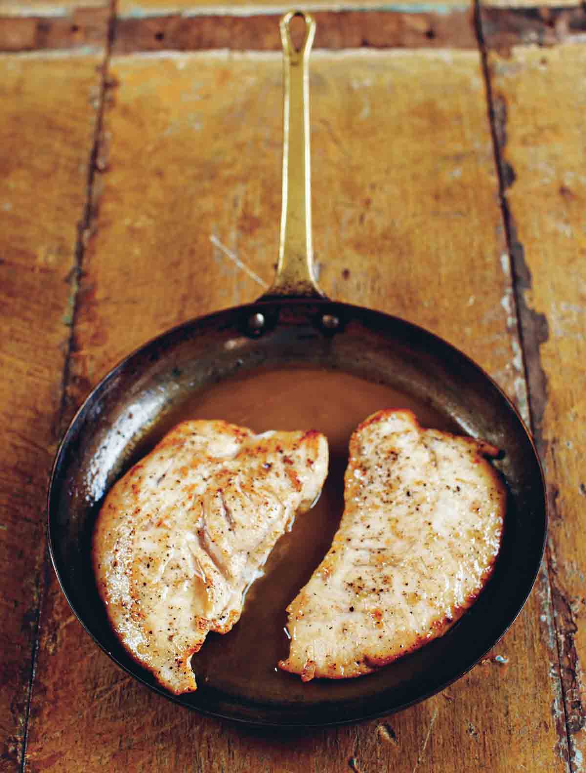 Two turkey cutlets with Marsala sauce in a skillet on a wooden table.