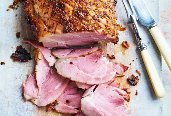 A glazed ham on a cutting board with several slices cut from the end and a carving fork and knife resting beside it.
