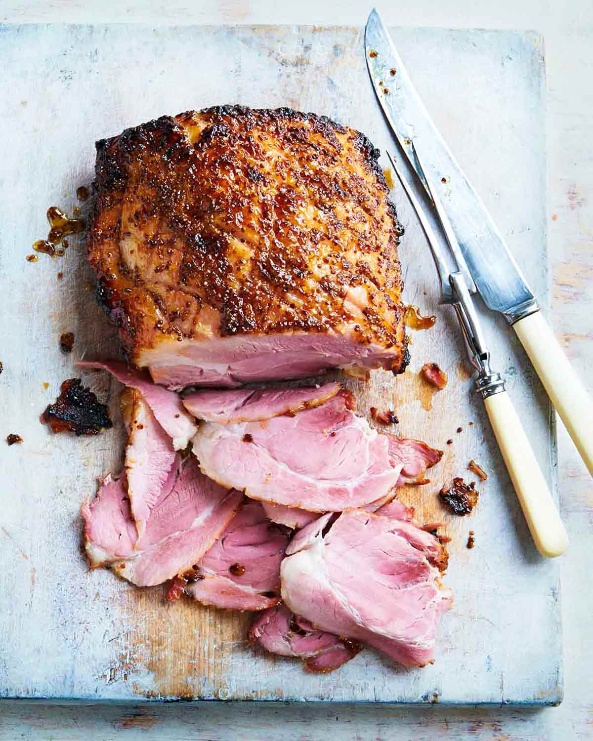 A glazed ham on a cutting board with several slices cut from the end and a carving fork and knife resting beside it.