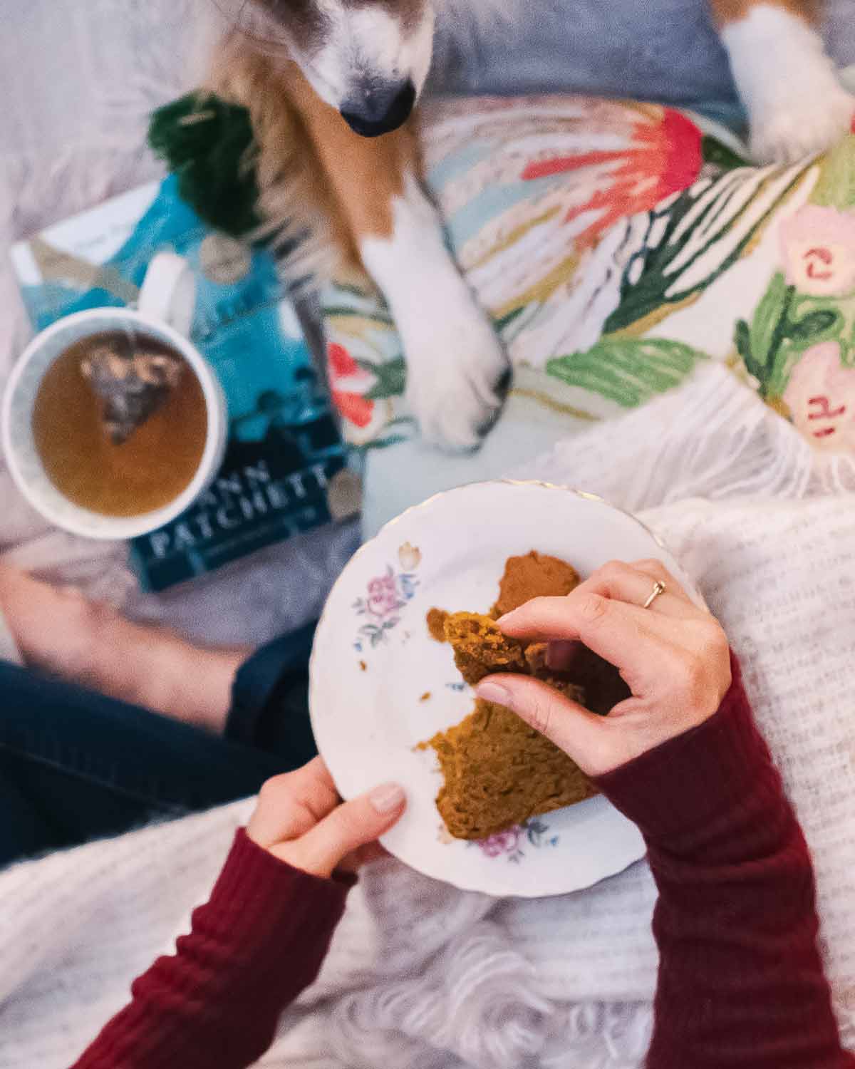 A woman eating a slice of pumpkin bread with a dog at her feet.