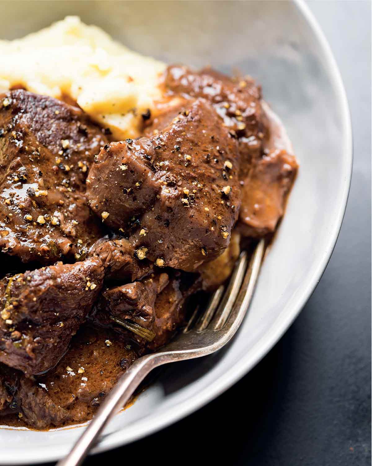 Pieces of beef stew with red wine and mashed potatoes in a white bowl with a fork nestled inside.