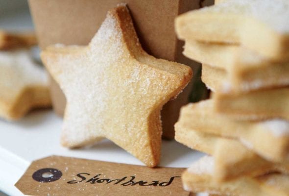 A stack of star-shaped cutout classic shortbread cookies, lightly dusted with sugar, and a brown takeout container and a gift tag