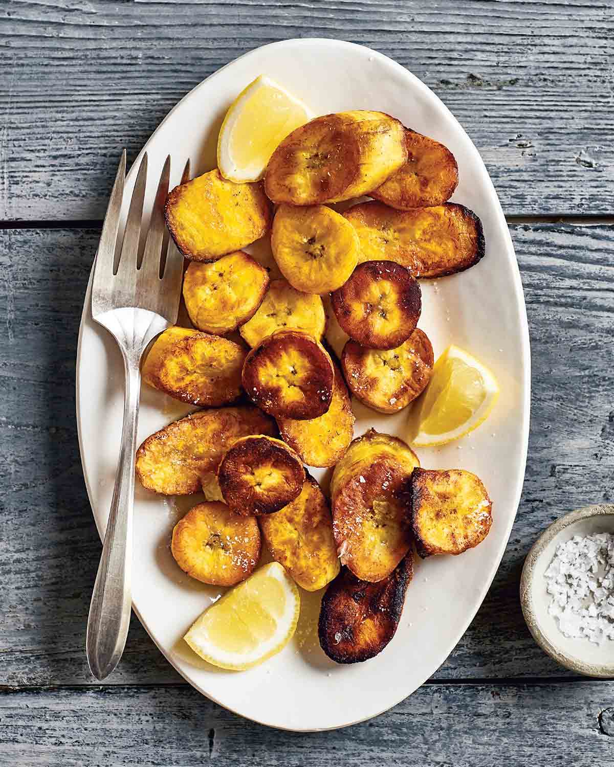 A white plate topped with slices of fried plantains or ndizi kaanga and lemon wedges with a fork on the side.