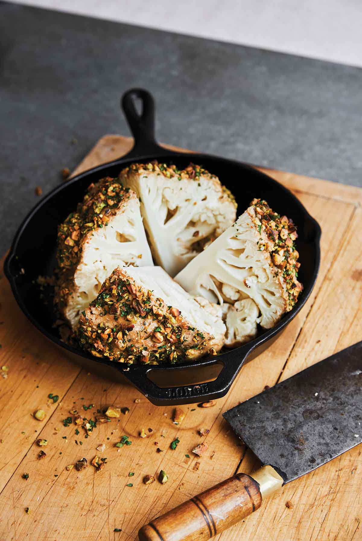 A whole roasted cauliflower with tahini and pistachios cut into four pieces in a cast-iron skillet.