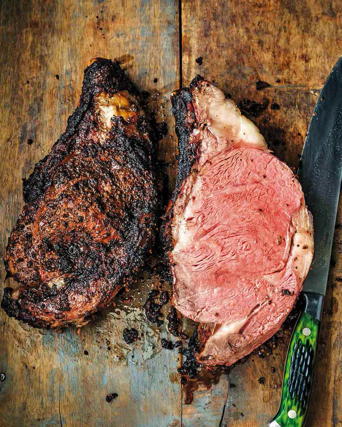 A smoked prime rib sliced in half on a wooden board with a green-handled knife beside it.