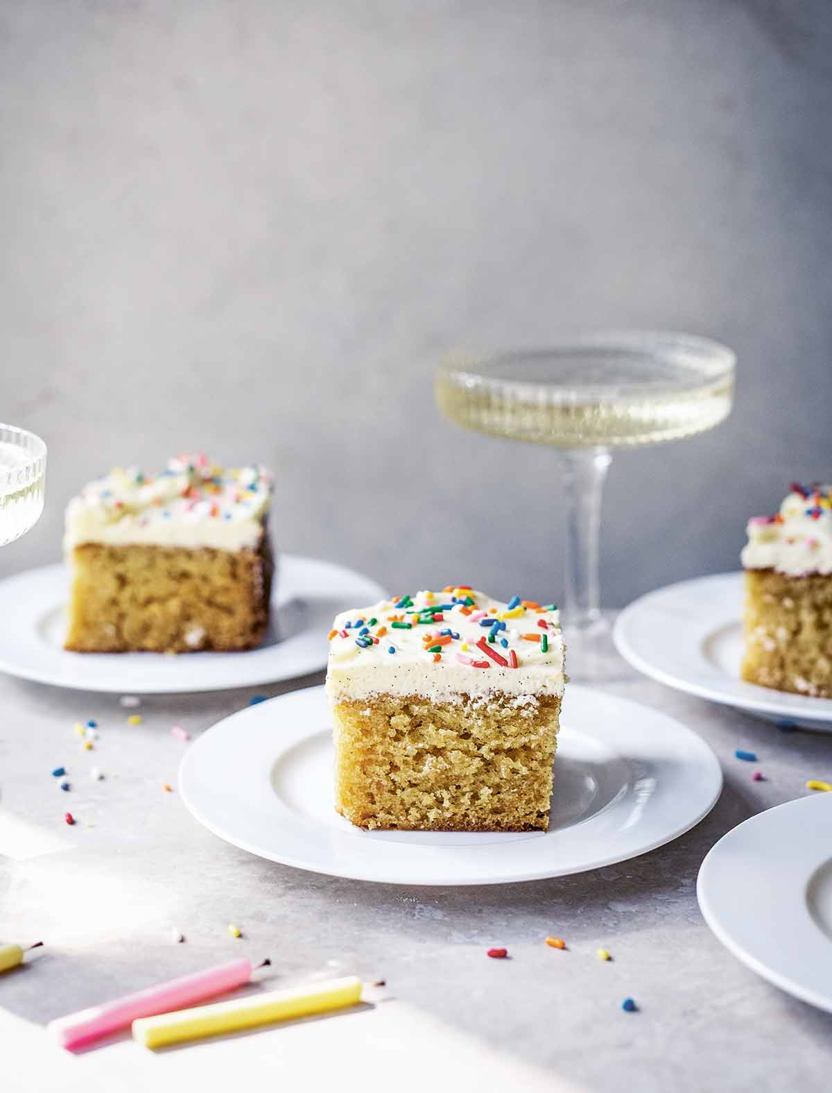 Squares of vanilla sheet cake with Swiss buttercream frosting and sprinkles on white plates with a coupe of Champagne in the background.