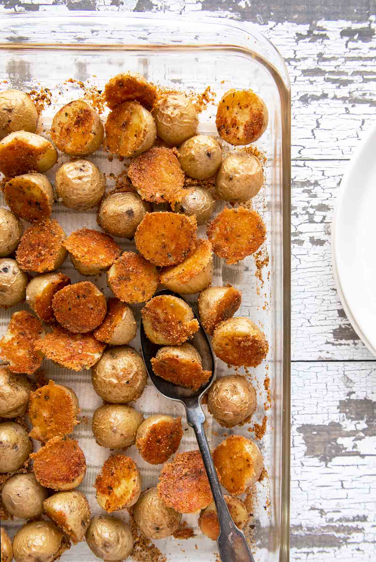 A baking dish filled with crispy herbed Parmesan potatoes on a white wooden surface with white plates beside it.