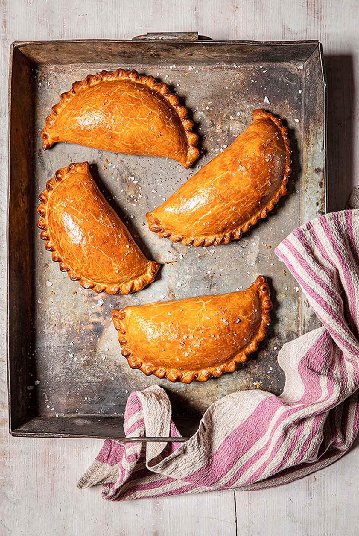 Four curried cauliflower and potato pasties in a metal baking dish with a pink and white striped towel on the edge.