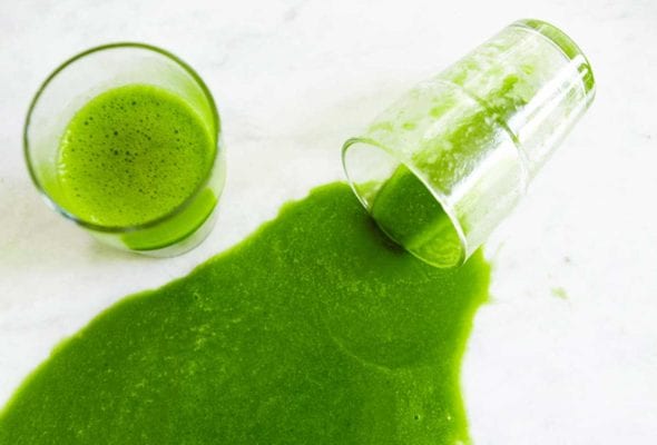 Two glasses of green smoothie, one of them tipped over and spilled with two spinach leaves beside it.