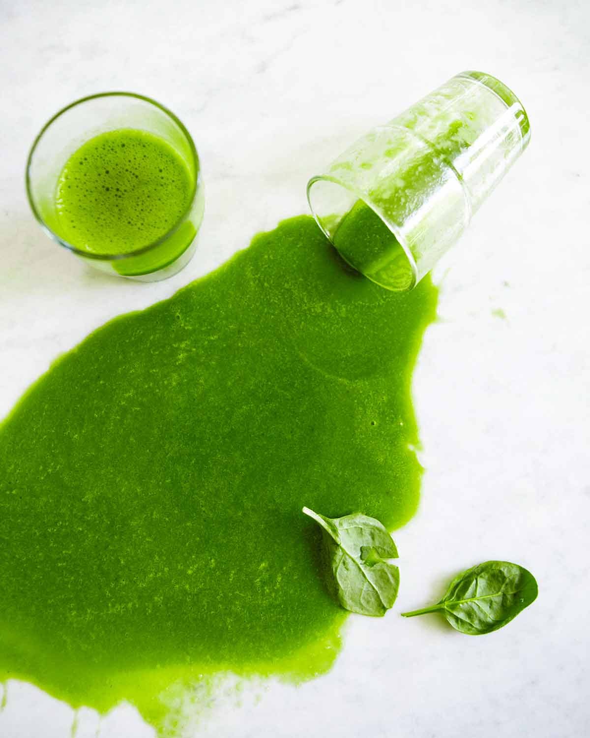 Two glasses of green smoothie, one of them tipped over and spilled with two spinach leaves beside it.