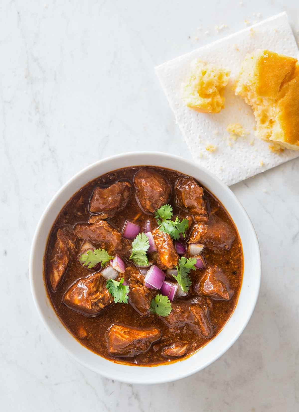 A white bowl filled with New Mexican red pork chili, garnished with red onion and cilantro.
