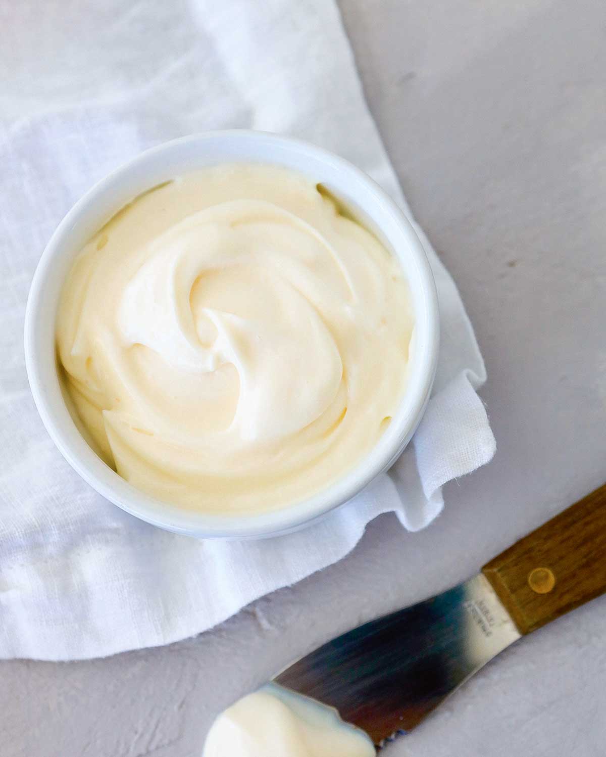A small dish filled with one minute mayo on a white cloth with a knife lying beside it.