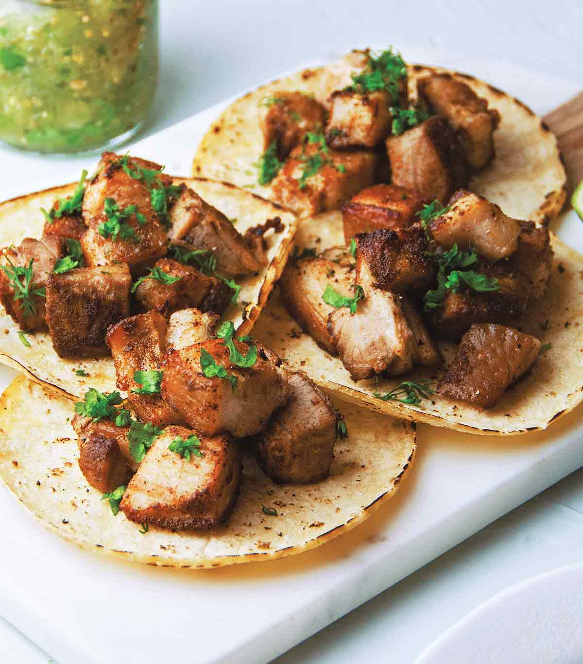 Four pork belly tacos garnished with cilantro and a jar of salsa in the background.
