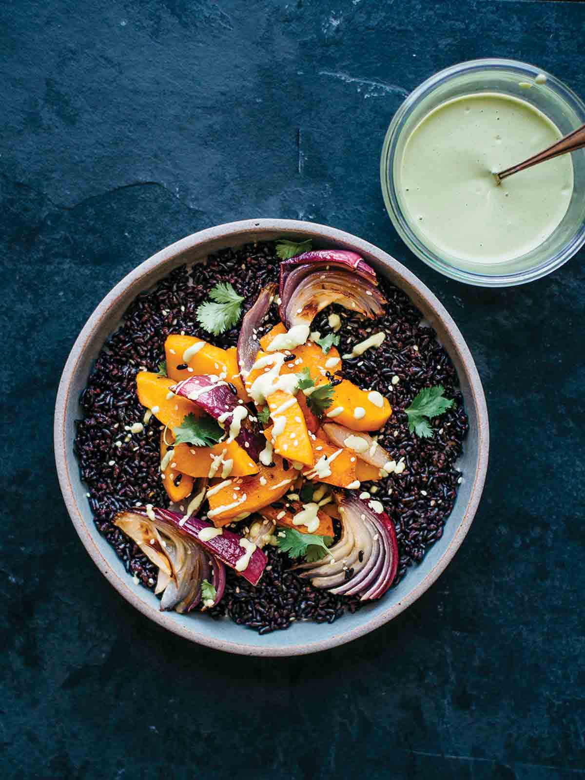 A white bowl filled with roasted kabocha squash, black rice, and sesame dressing drizzled over and in a glass on the side.