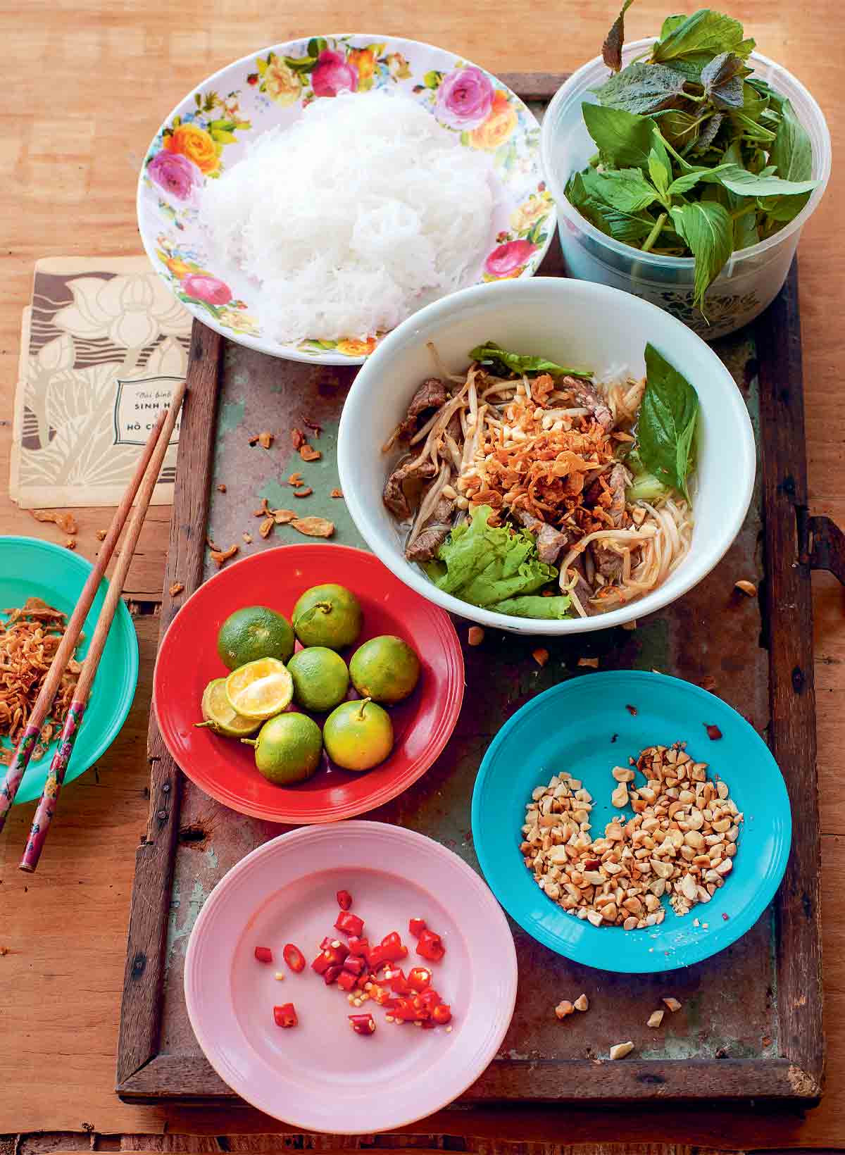 A white bowl of Vietnamese bun bo with bowls of lime, peanuts, chili, basil, and vermicelli noodles on the side.