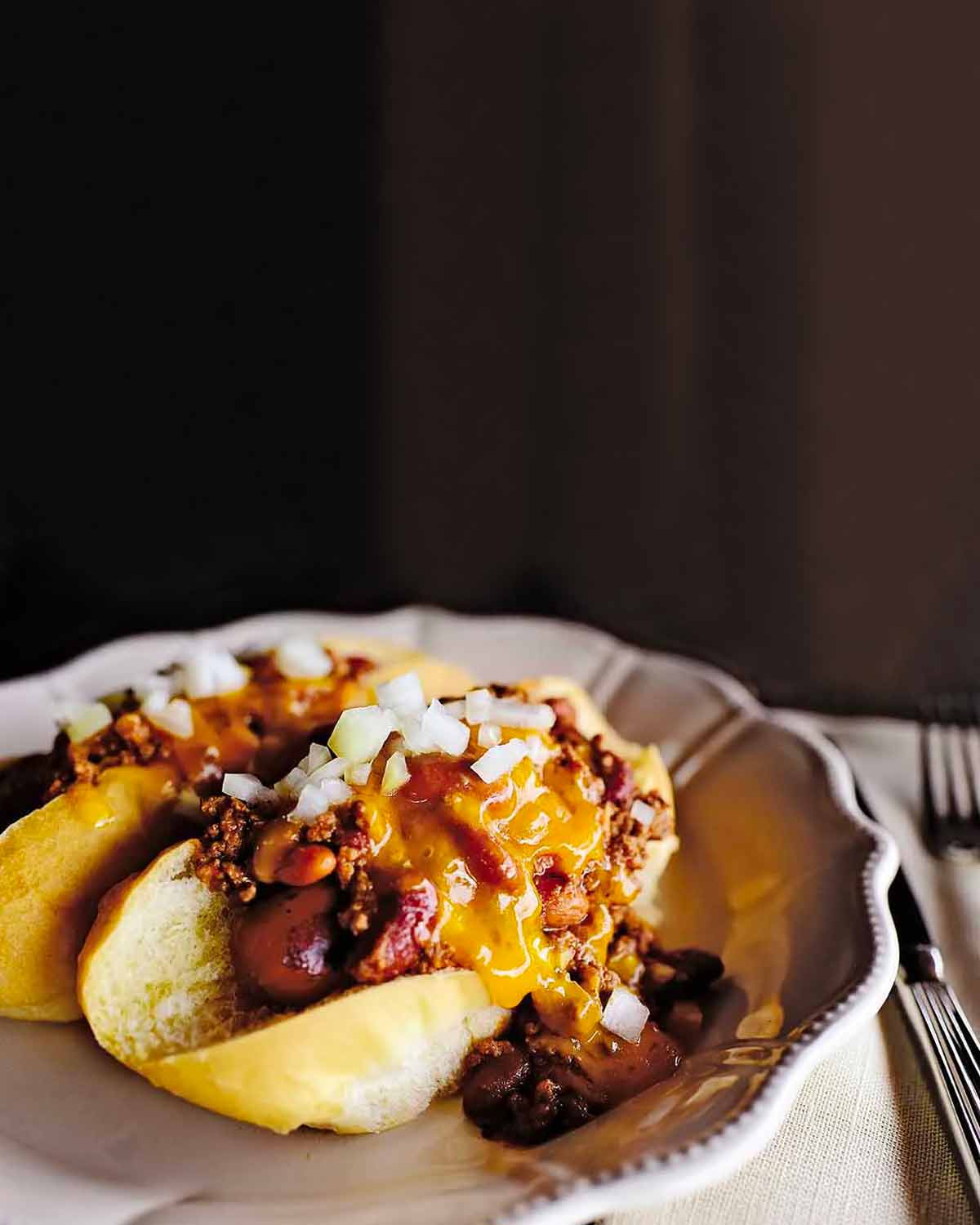 Two chili dogs on a white plate with a fork and knife on the side.