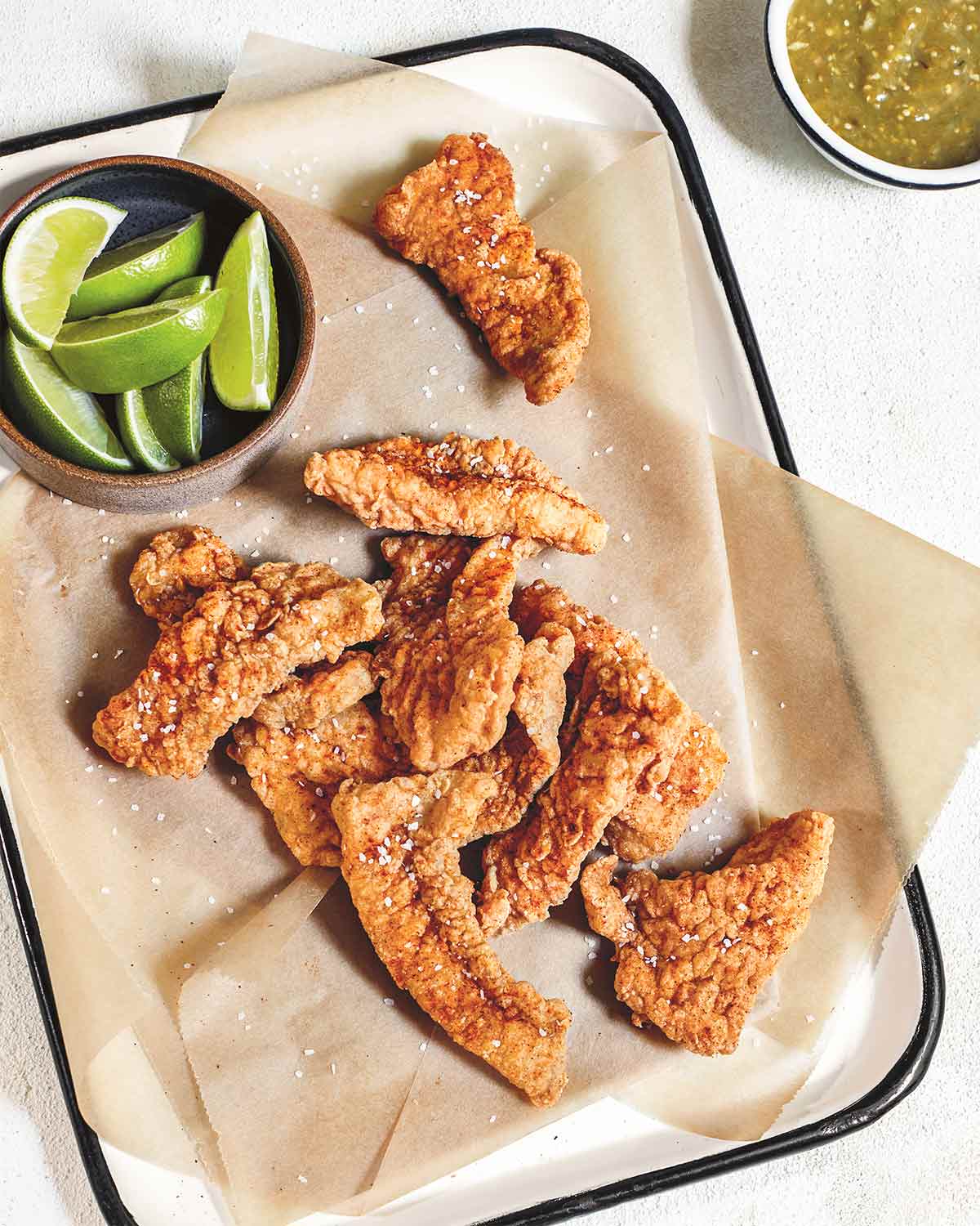 Pieces of fried fish with masa on a parchment-lined tray with a dish of limes on the tray and a dish of salsa on the side.